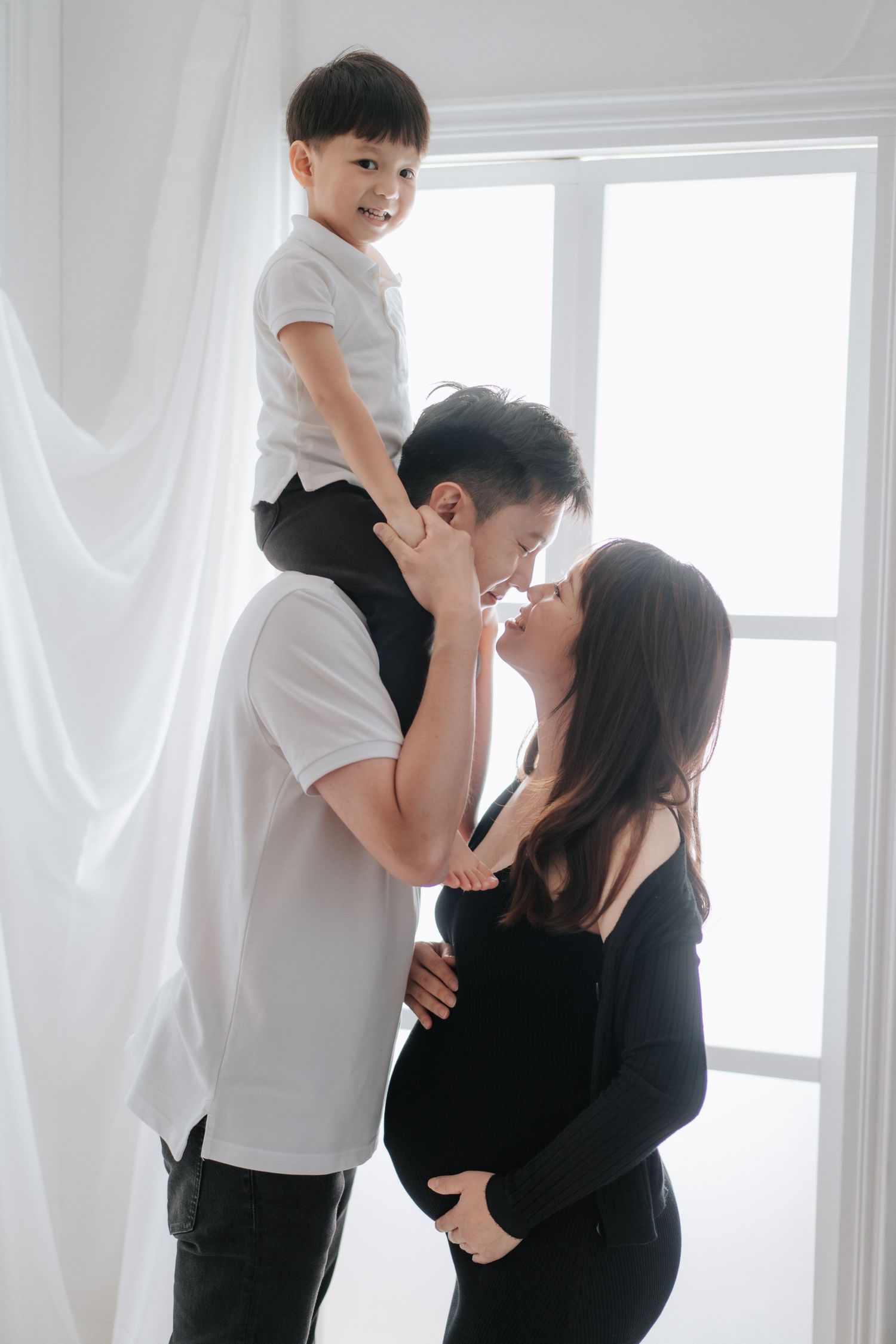 A person in black attire sits on a white bed near sheer curtains in a bright, minimalist setting.