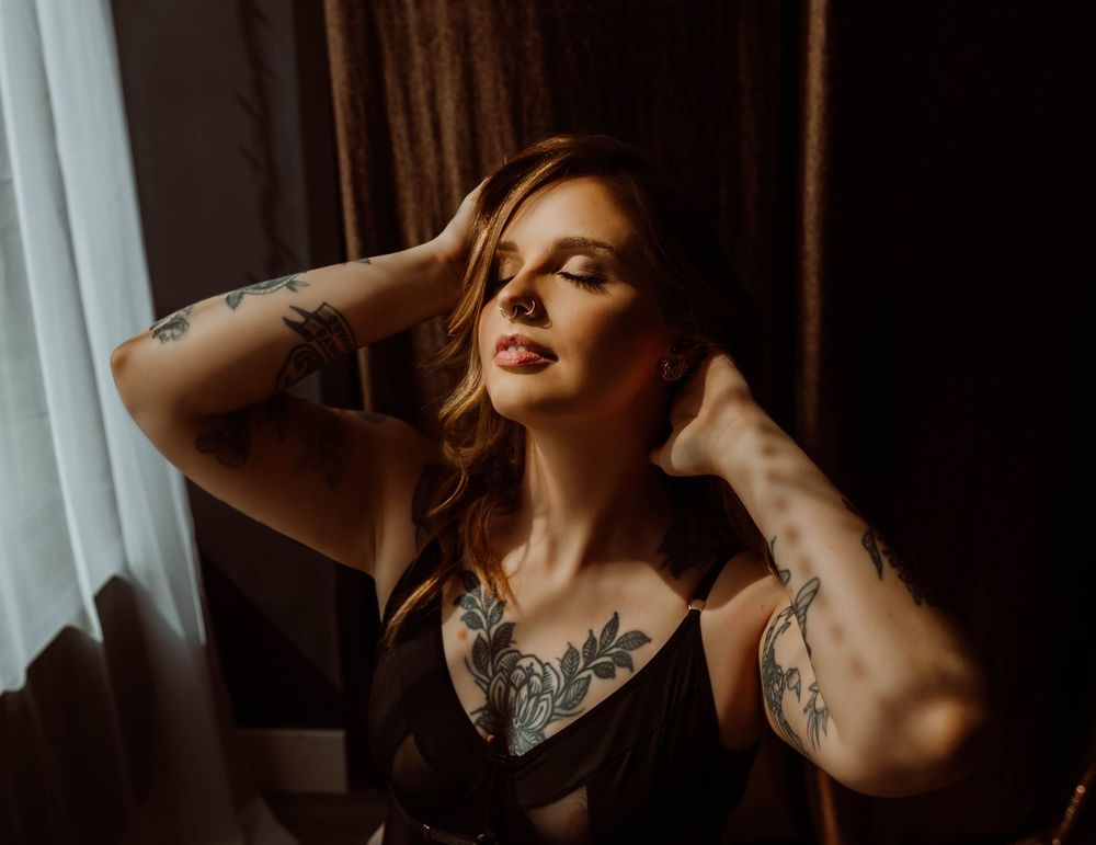 A woman in a black tank top with tattoos poses in a moody, dark interior setting with dramatic lighting with Boudoir by KJ.