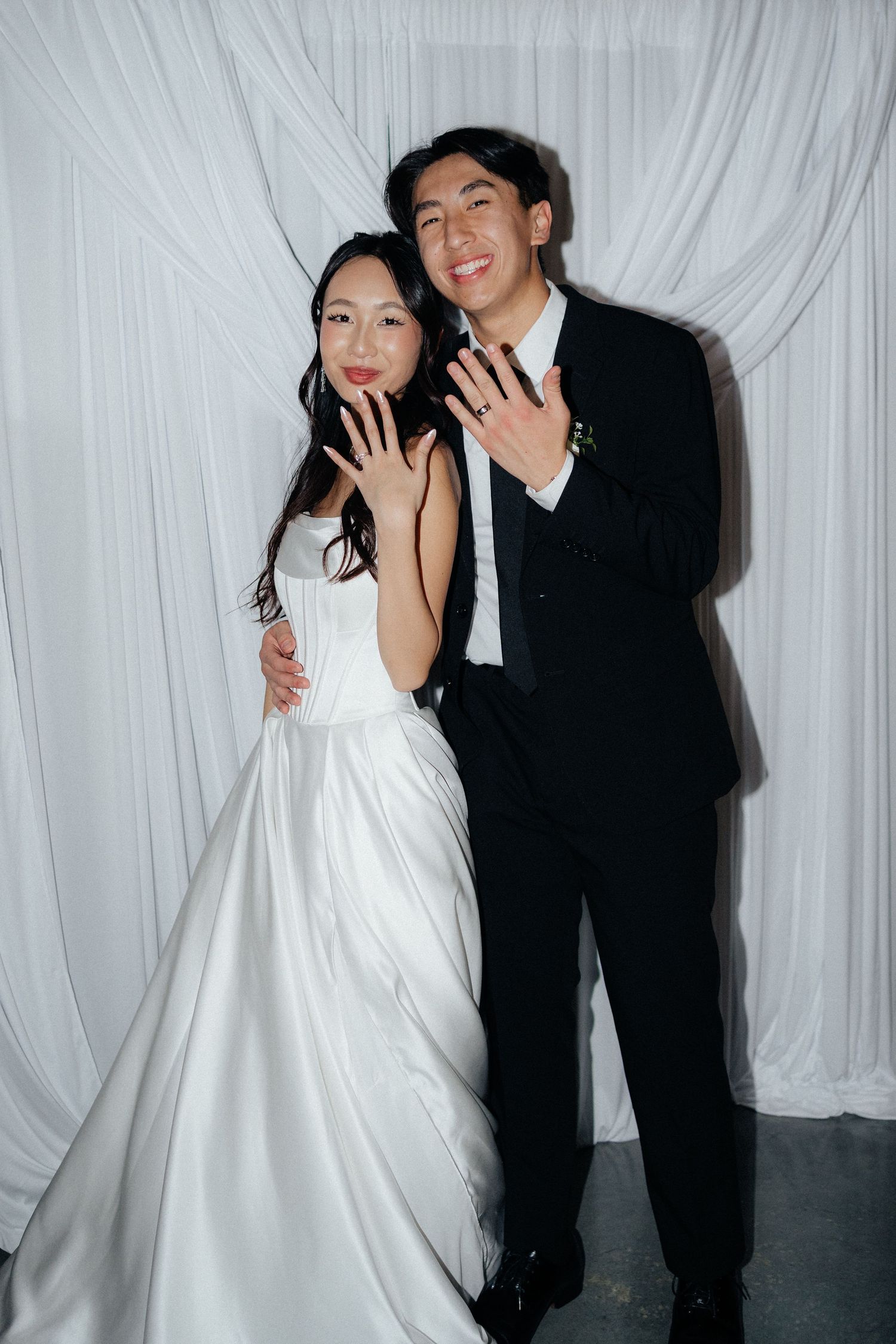 A couple in formal wedding attire poses playfully against a white draped backdrop at their reception.