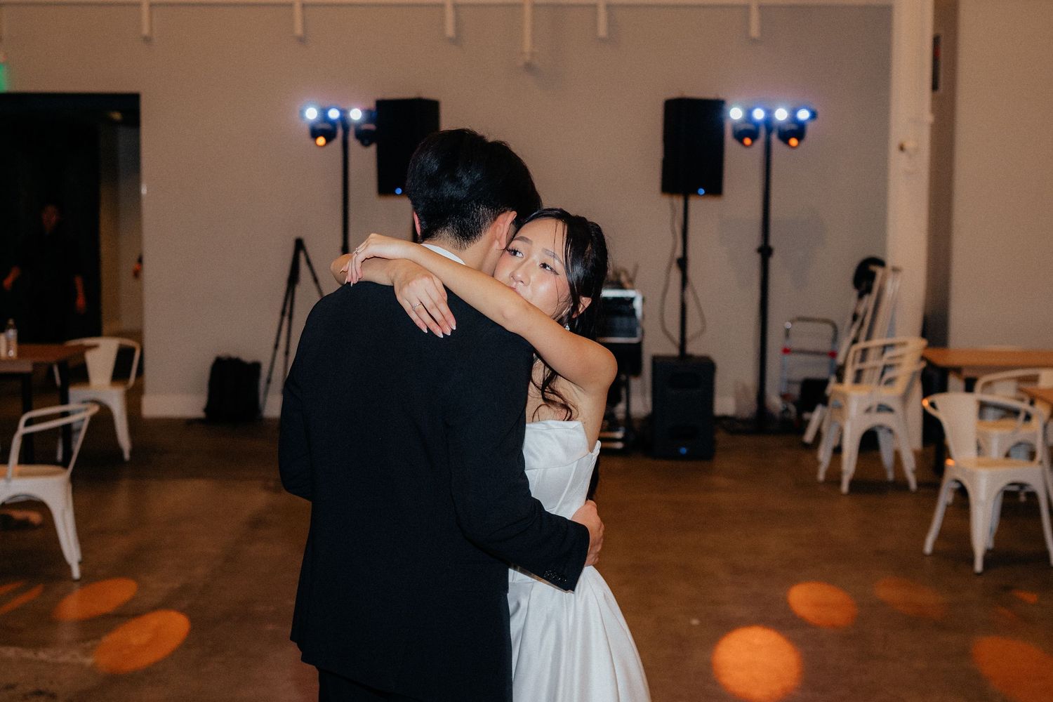 A couple dances together during their first dance at a wedding reception with white chairs and blue lighting in the background.