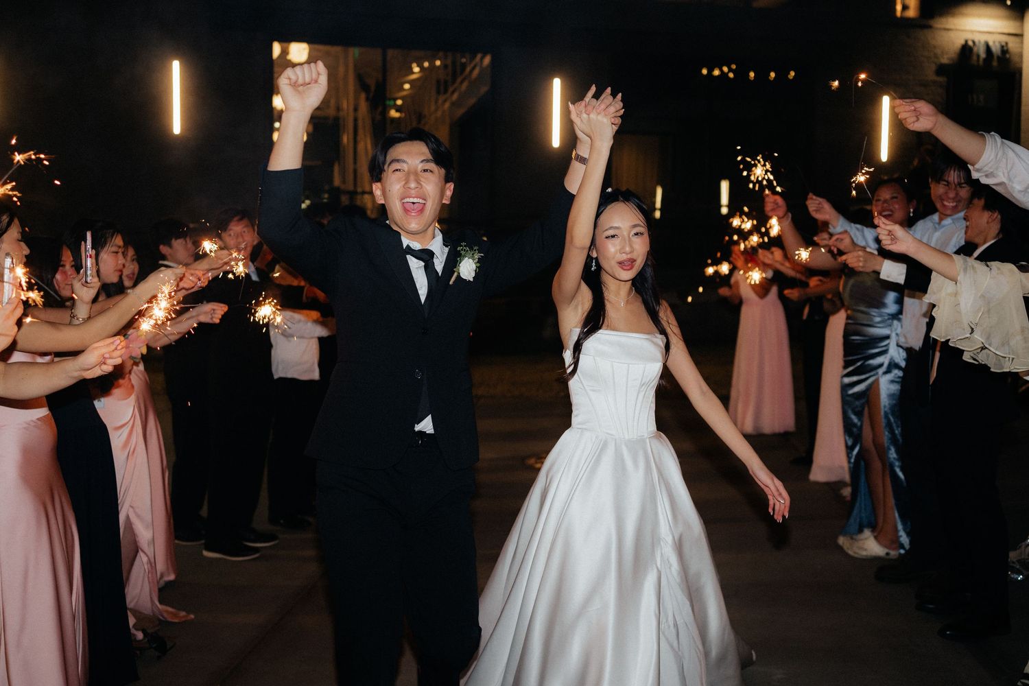 Wedding party dances joyfully during evening reception with string lights overhead.
