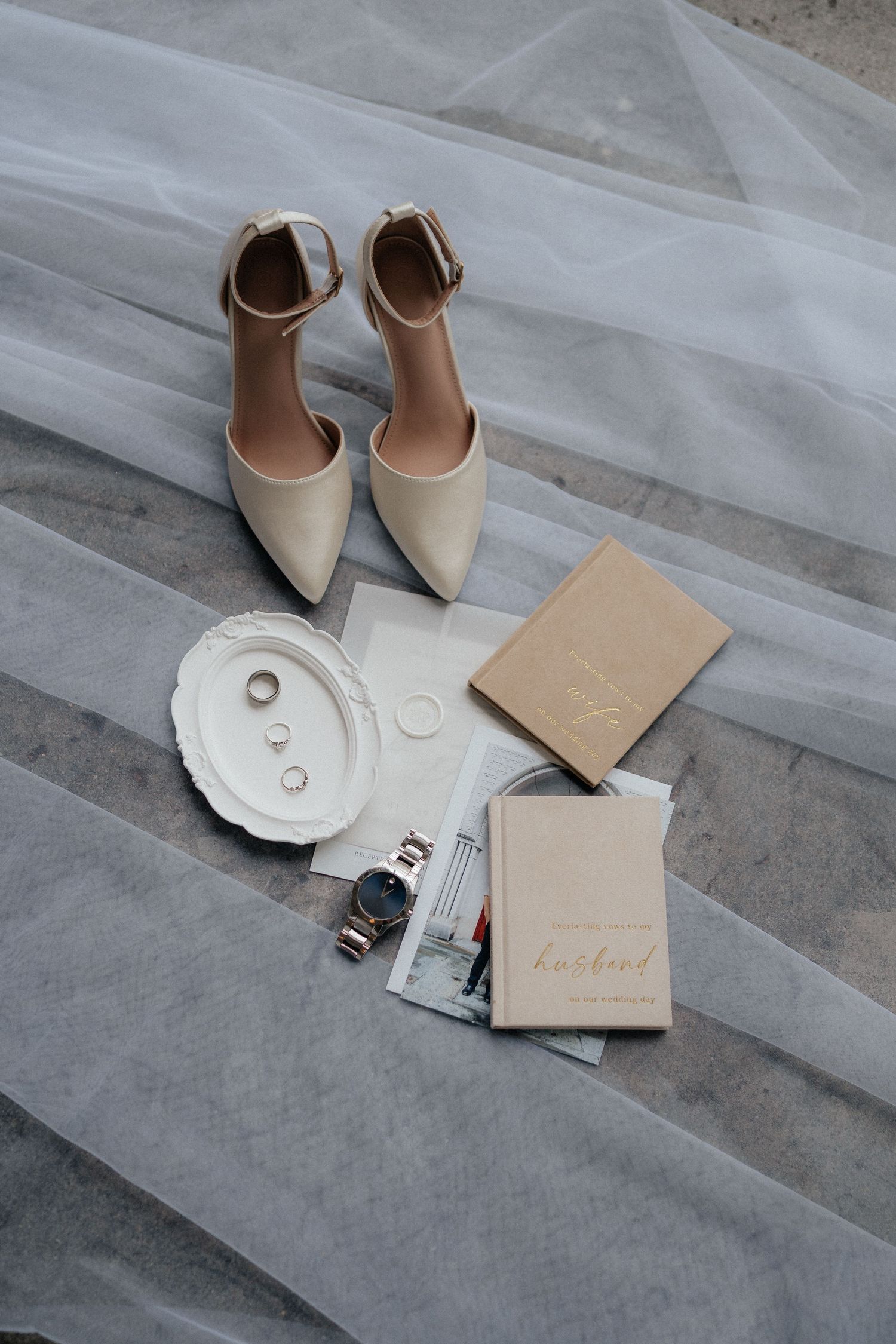 Wedding accessories including beige heels and invitation cards arranged on grey fabric.