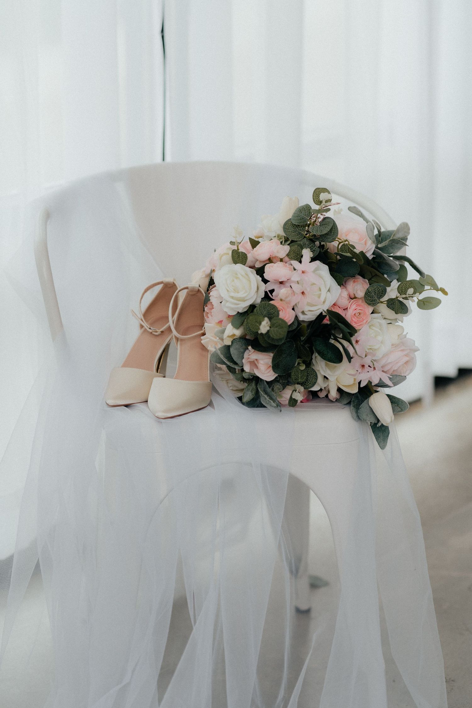 Elegant bridal accessories featuring blush pink heels and a delicate bouquet with pink roses against white tulle fabric.
