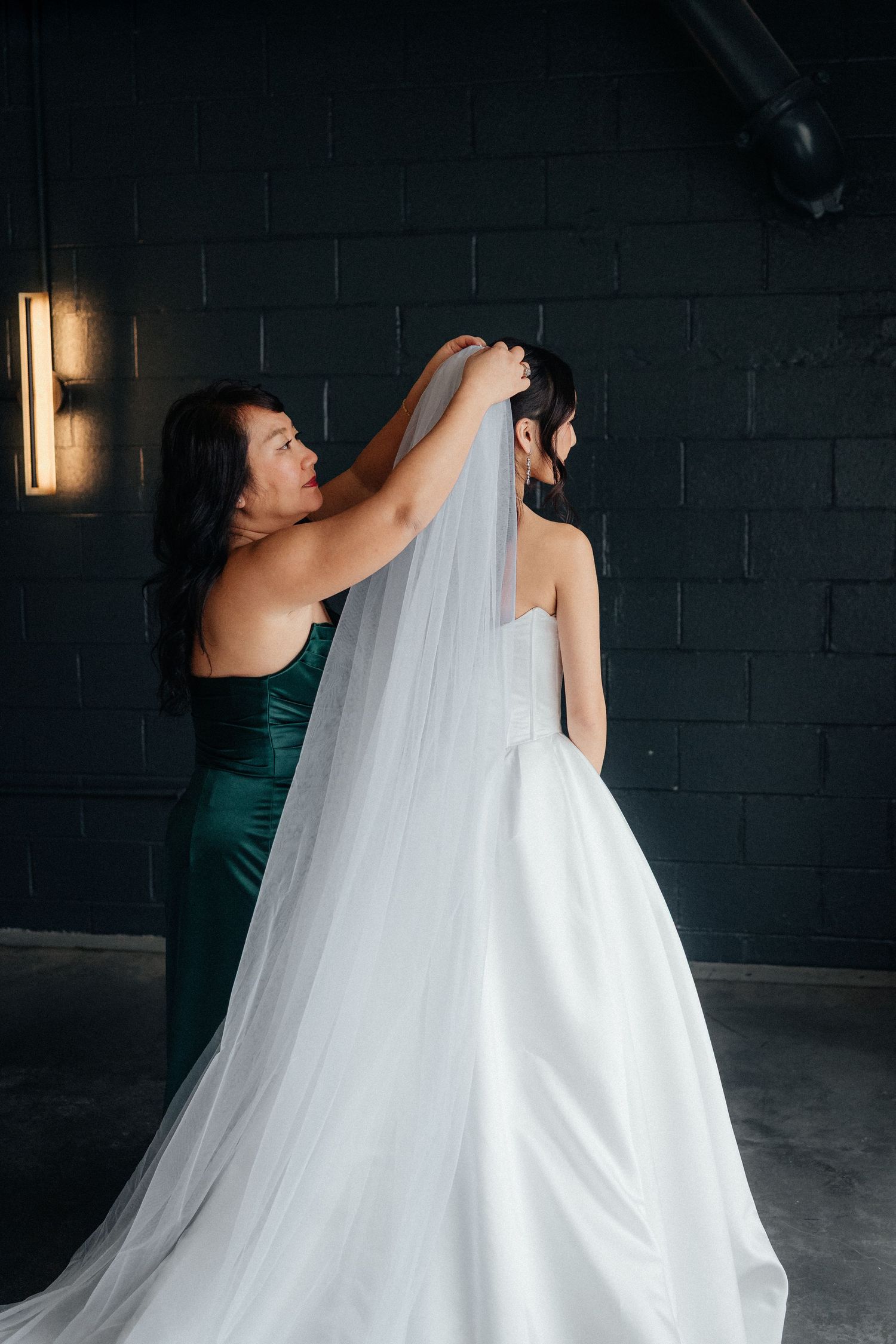 Bridesmaids in dark green dresses help adjust a flowing white wedding veil in a dramatic indoor setting.