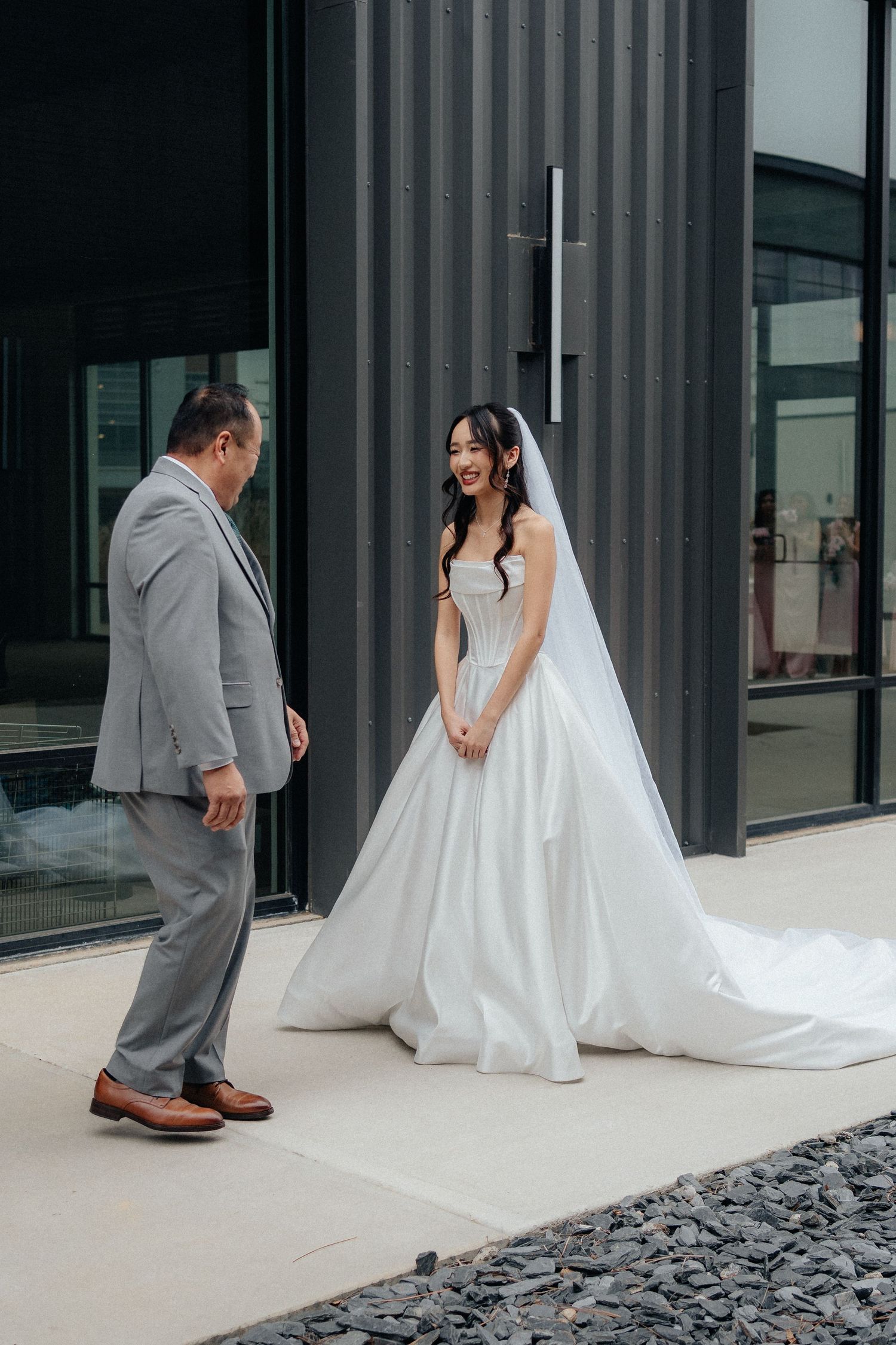 A bride in a flowing white gown and groom in gray suit share a tender first look moment against a modern building exterior.