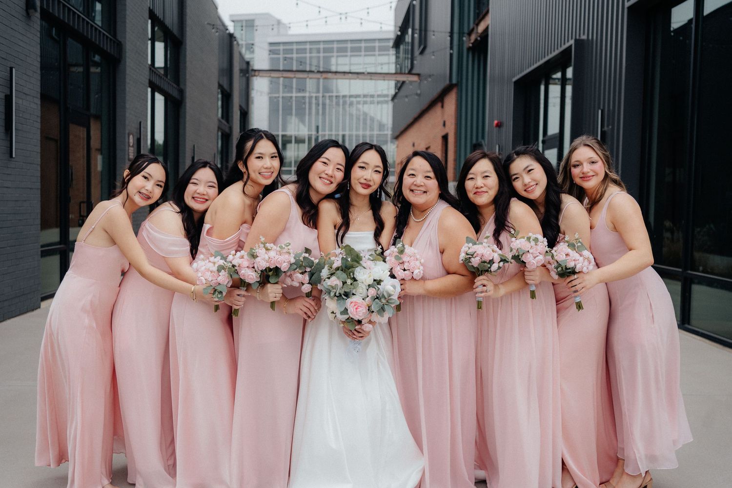Bridesmaids in matching pink dresses pose together holding white floral bouquets at a modern wedding venue.