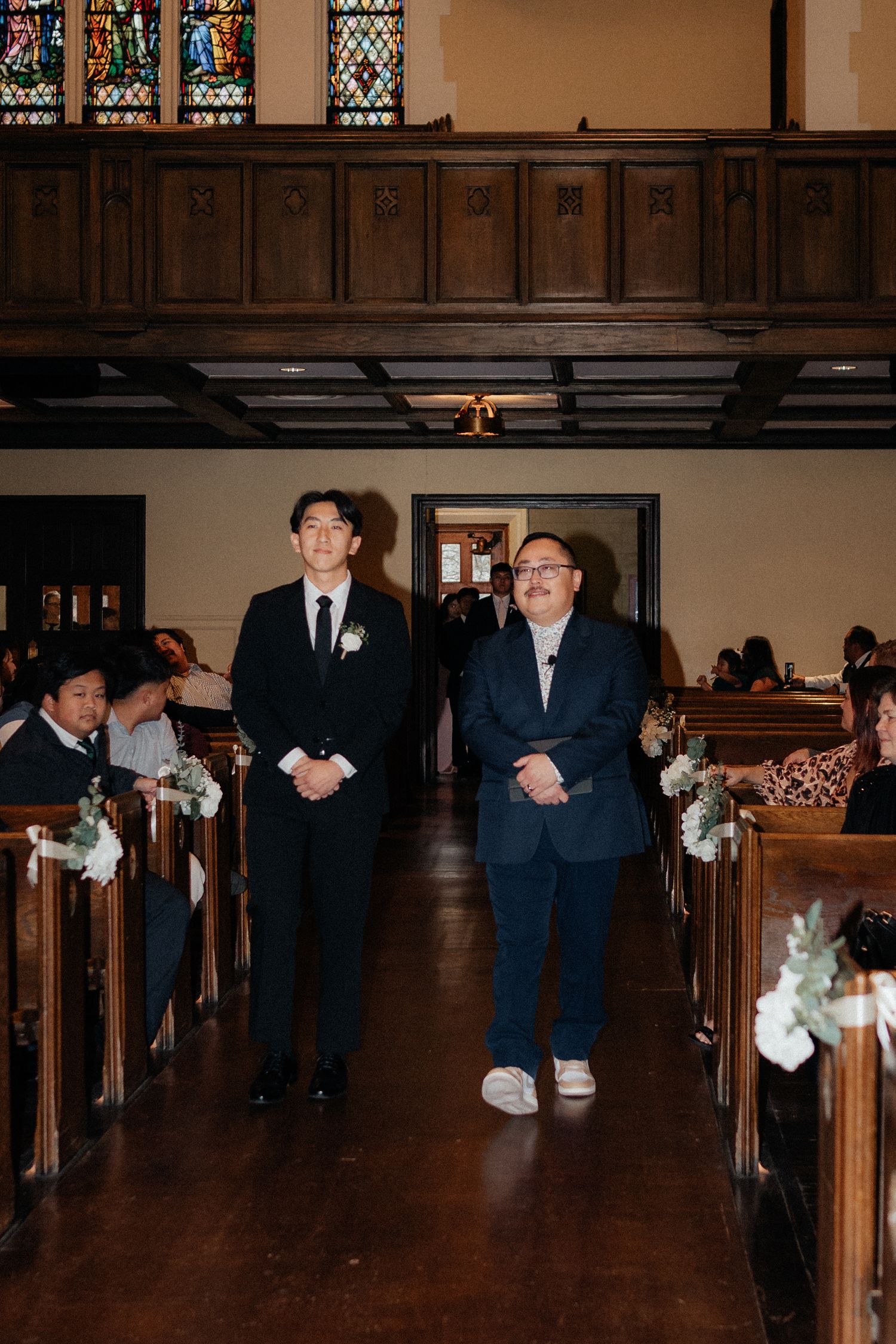 Wedding party members in navy suits walking down the aisle of a church with stained glass windows.