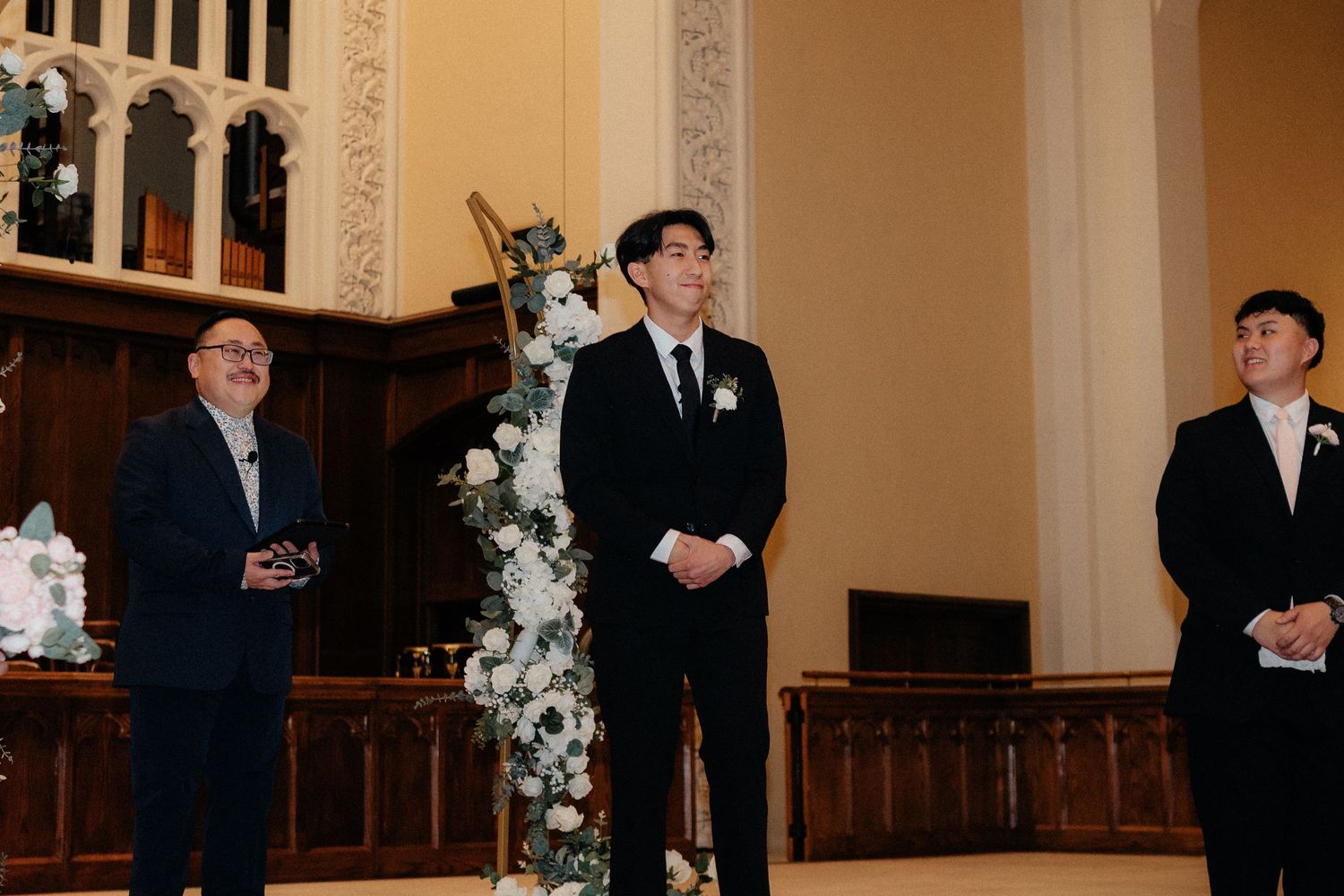 Three people stand at the altar of a church with elegant white floral decorations during a wedding ceremony.