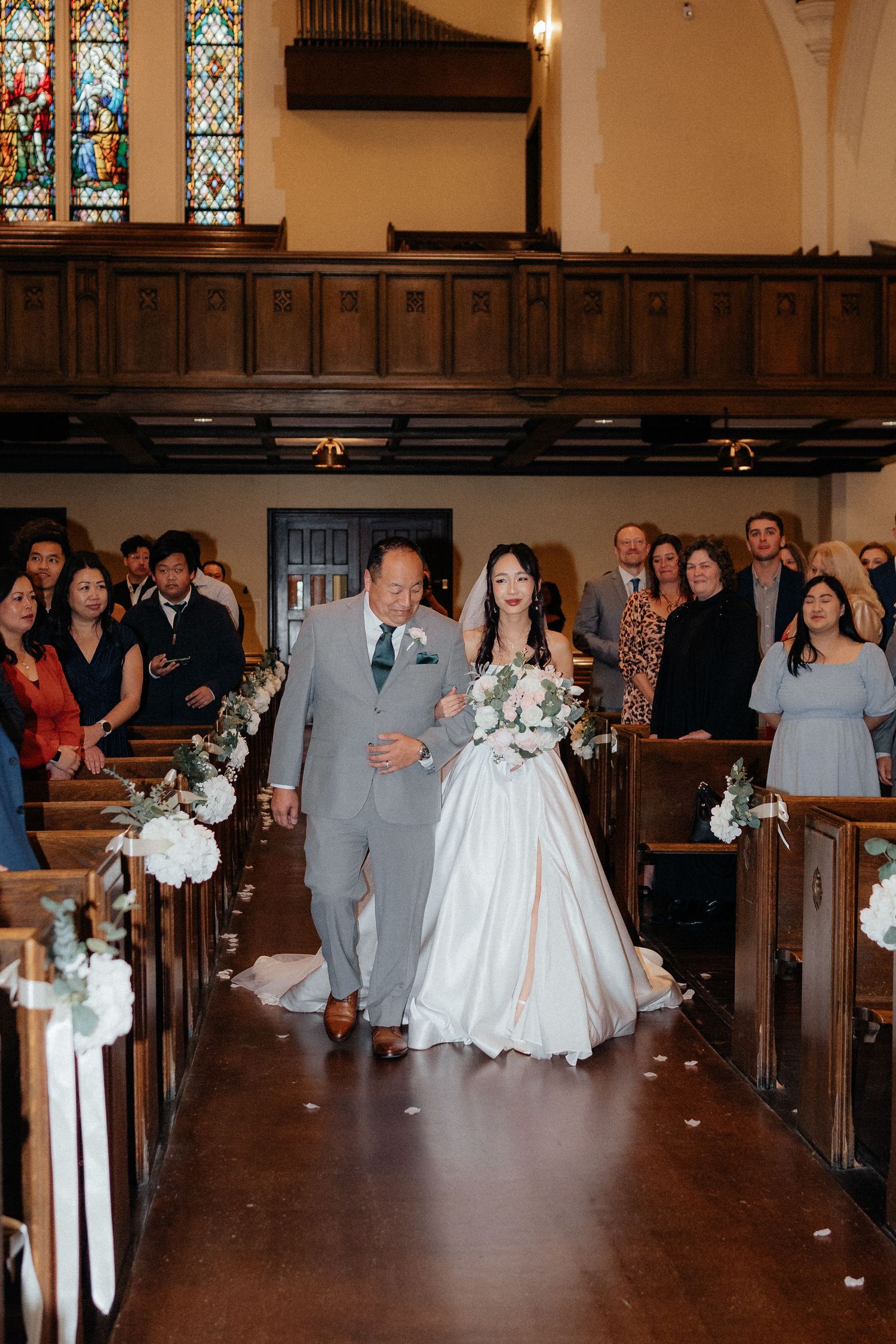 Wedding ceremony procession in a traditional church with wooden pews decorated with floral arrangements.