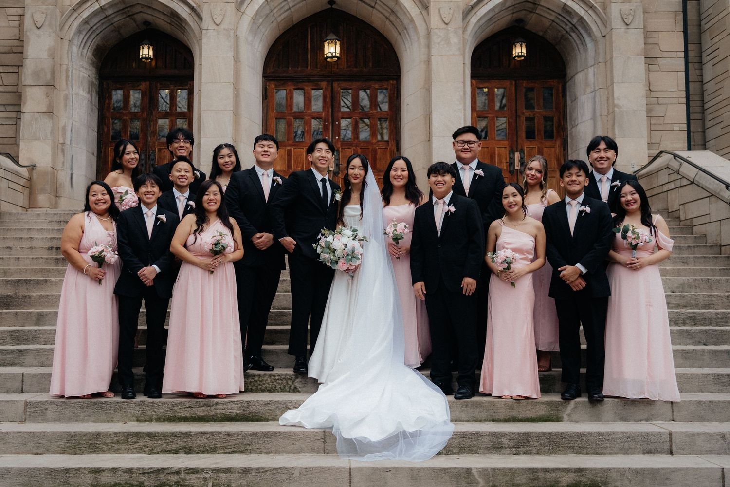 Wedding party poses on grand stone church steps with bridesmaids in pink dresses and groomsmen in dark suits.