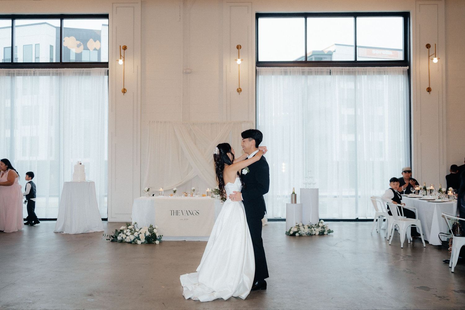 A couple dances in an elegant white studio space with large windows and natural light streaming in.