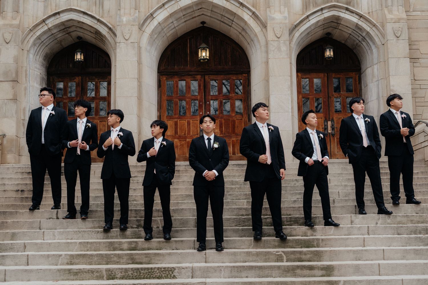 Wedding party in formal attire poses on stone church steps for group photo.