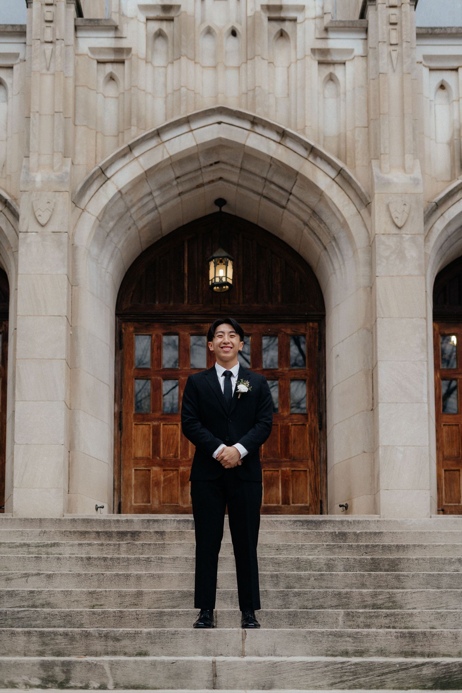 A person in a black suit stands alone on grand stone steps in front of arched wooden doors of a historic building.