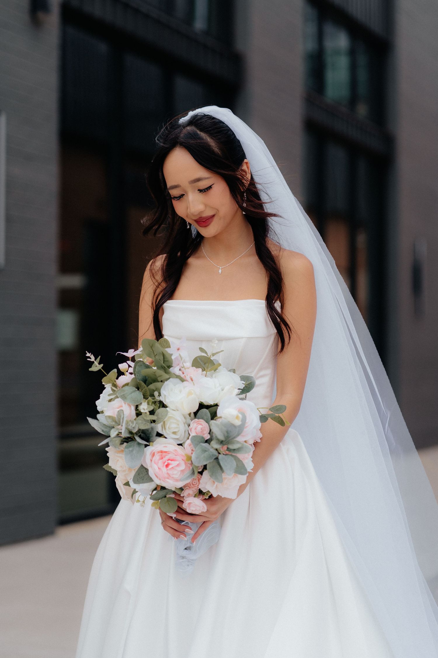 A bride in a white strapless wedding gown holds a pastel bouquet of roses and eucalyptus against an urban backdrop.
