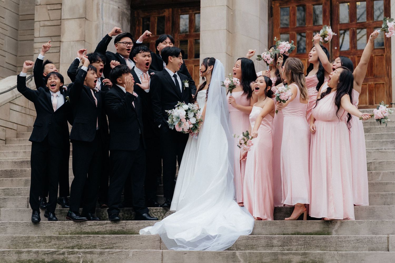 Wedding party in pink dresses and dark suits posing together on stone church steps.