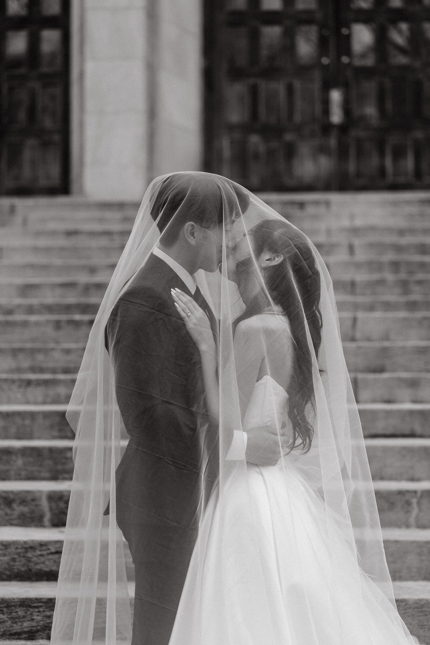 A romantic black and white wedding photo shows a couple embracing under a flowing white veil on stone steps.
