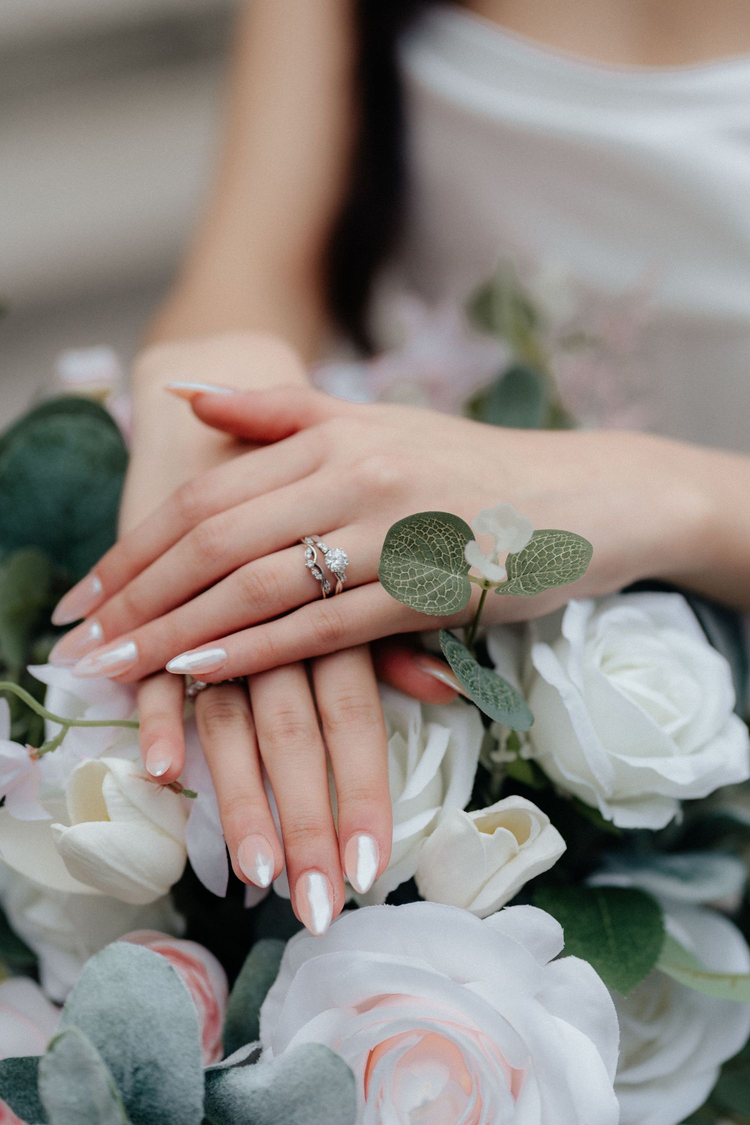 Close-up of manicured hands with engagement ring resting on white roses and green foliage.