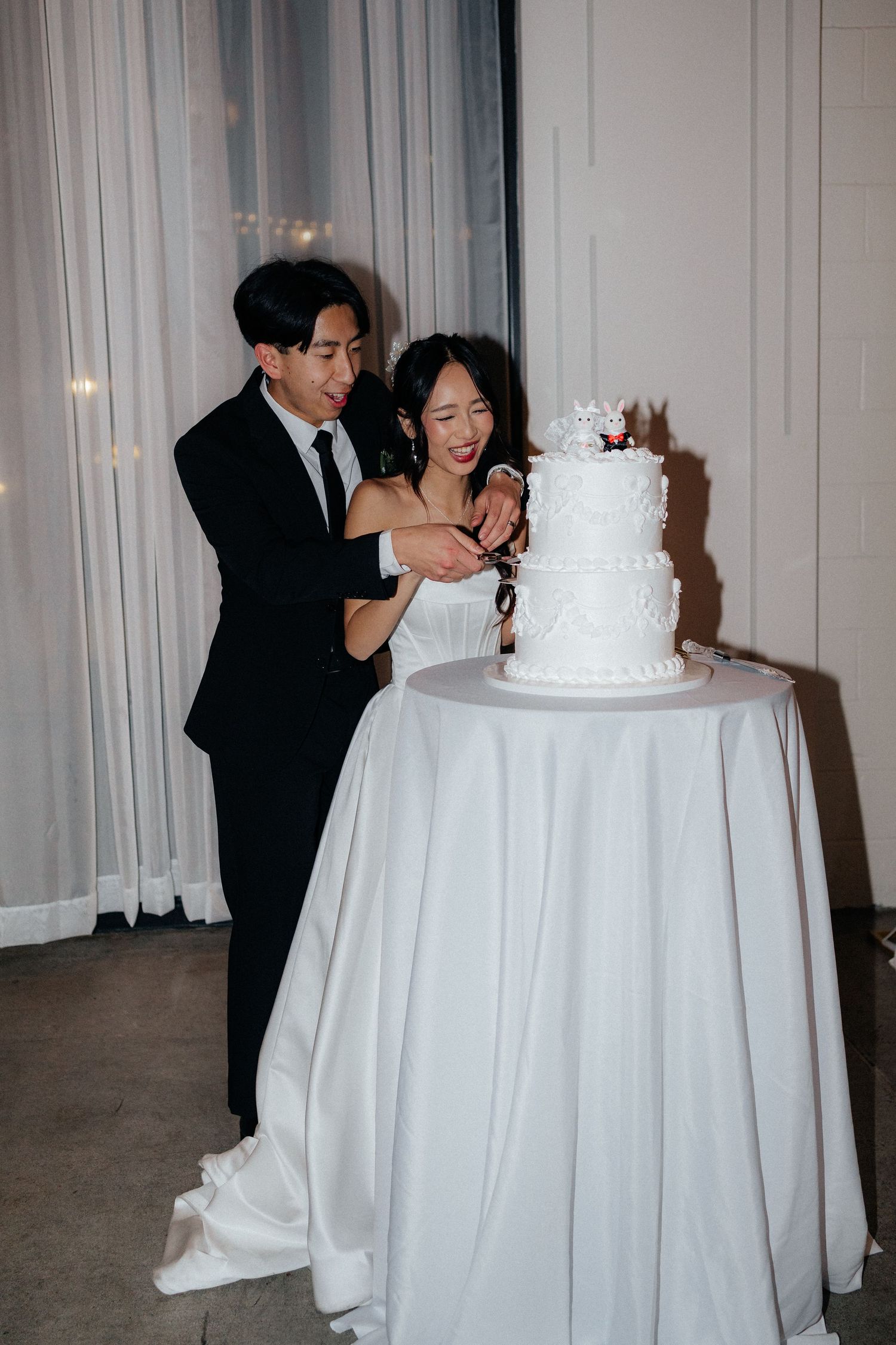 Wedding guests gather around a white tiered cake displayed on an elegant table with white tablecloth at a reception.