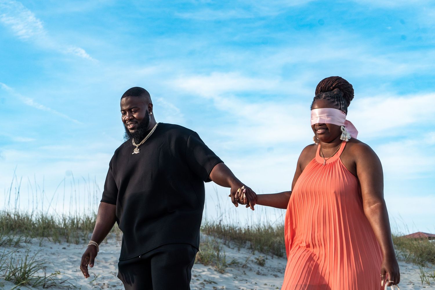 A couple walks together on a sandy beach path surrounded by sea grass and dunes under a bright blue sky.