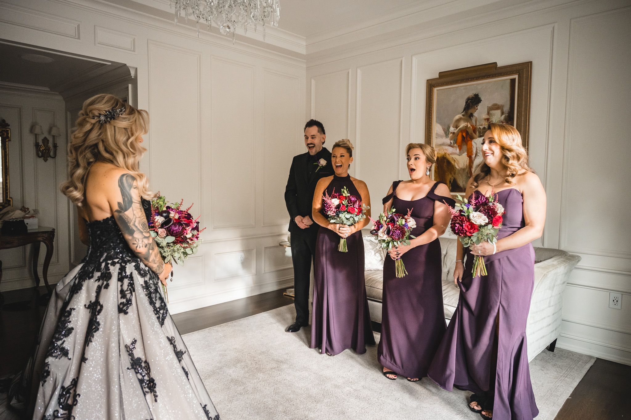 Wedding party members in purple dresses gather in an elegant white room with ornate trim and artwork.