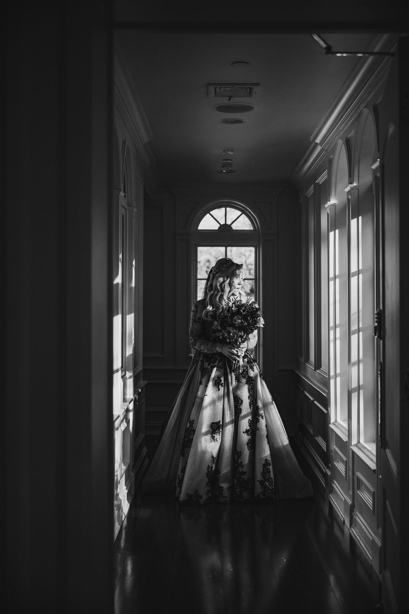 Dramatic black and white photo of a wedding dress silhouette in a sunlit hallway with arched window.