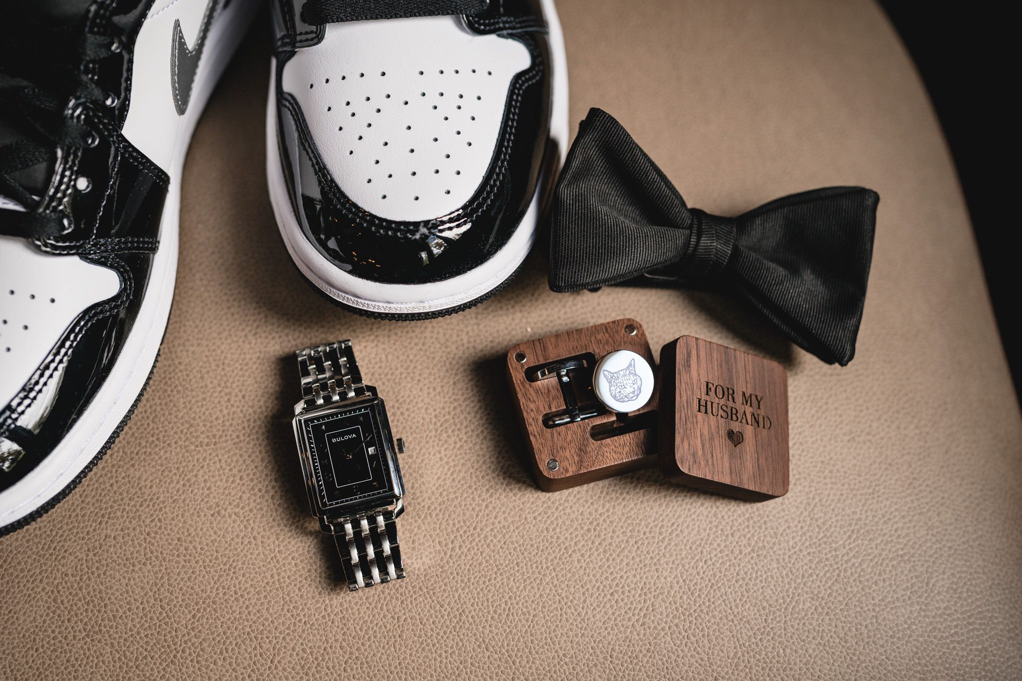 Wedding accessories including dress shoes, black bowtie, watch and wooden ring box on beige surface.