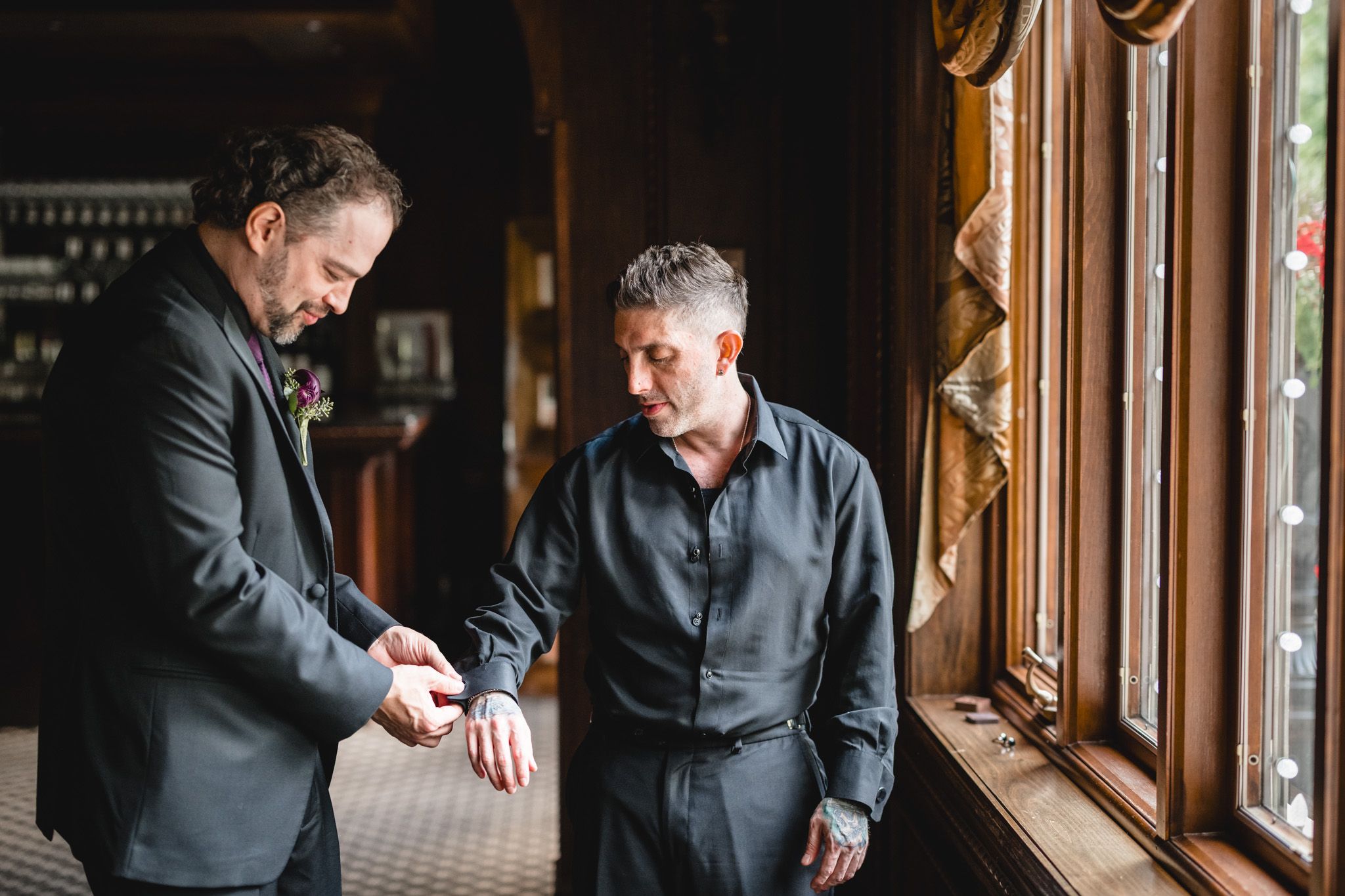 Two men in formal attire adjusting their cuffs by a window in a dimly lit room.