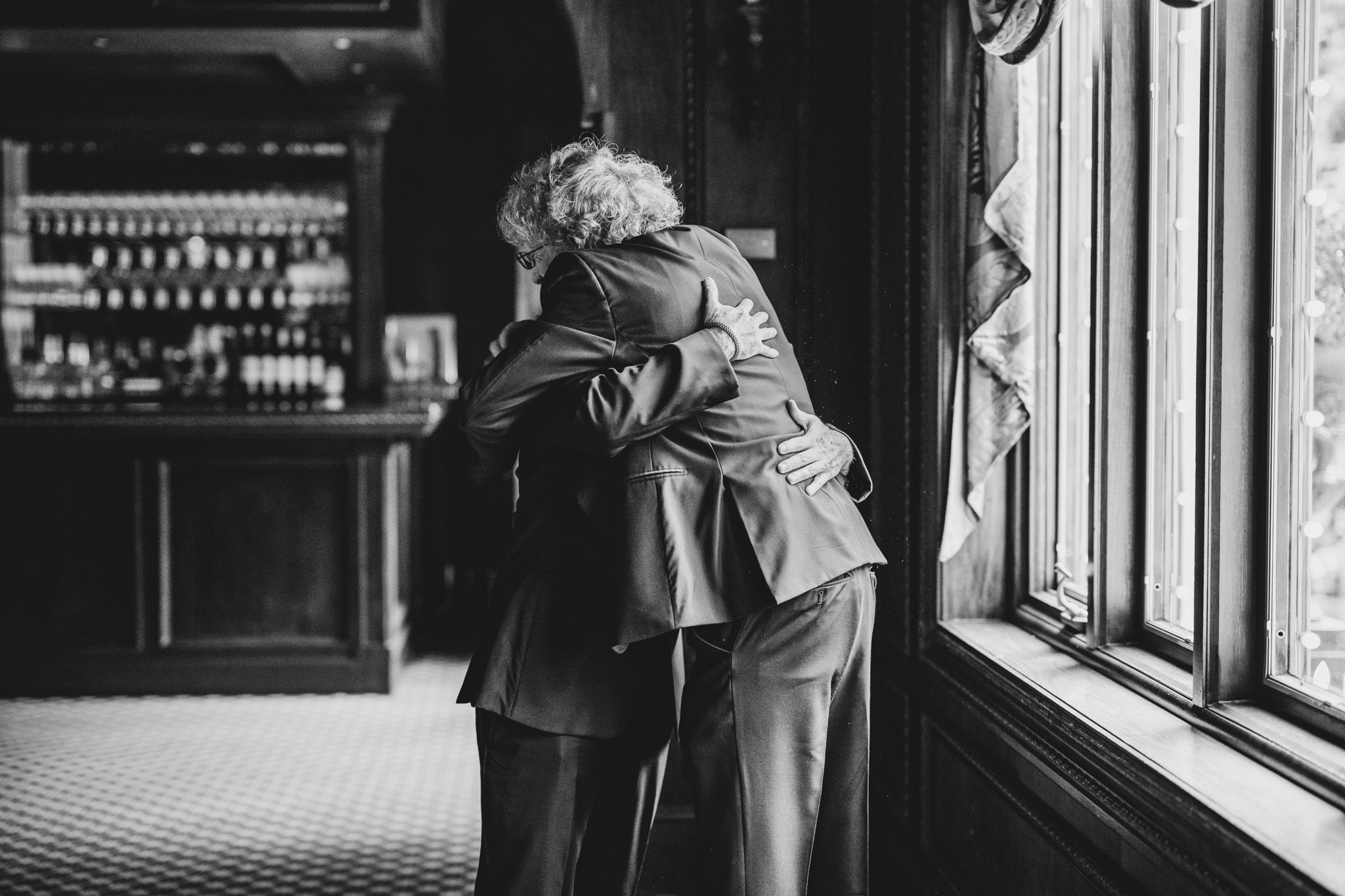 Emotional embrace captured in black and white near a window in formal attire.
