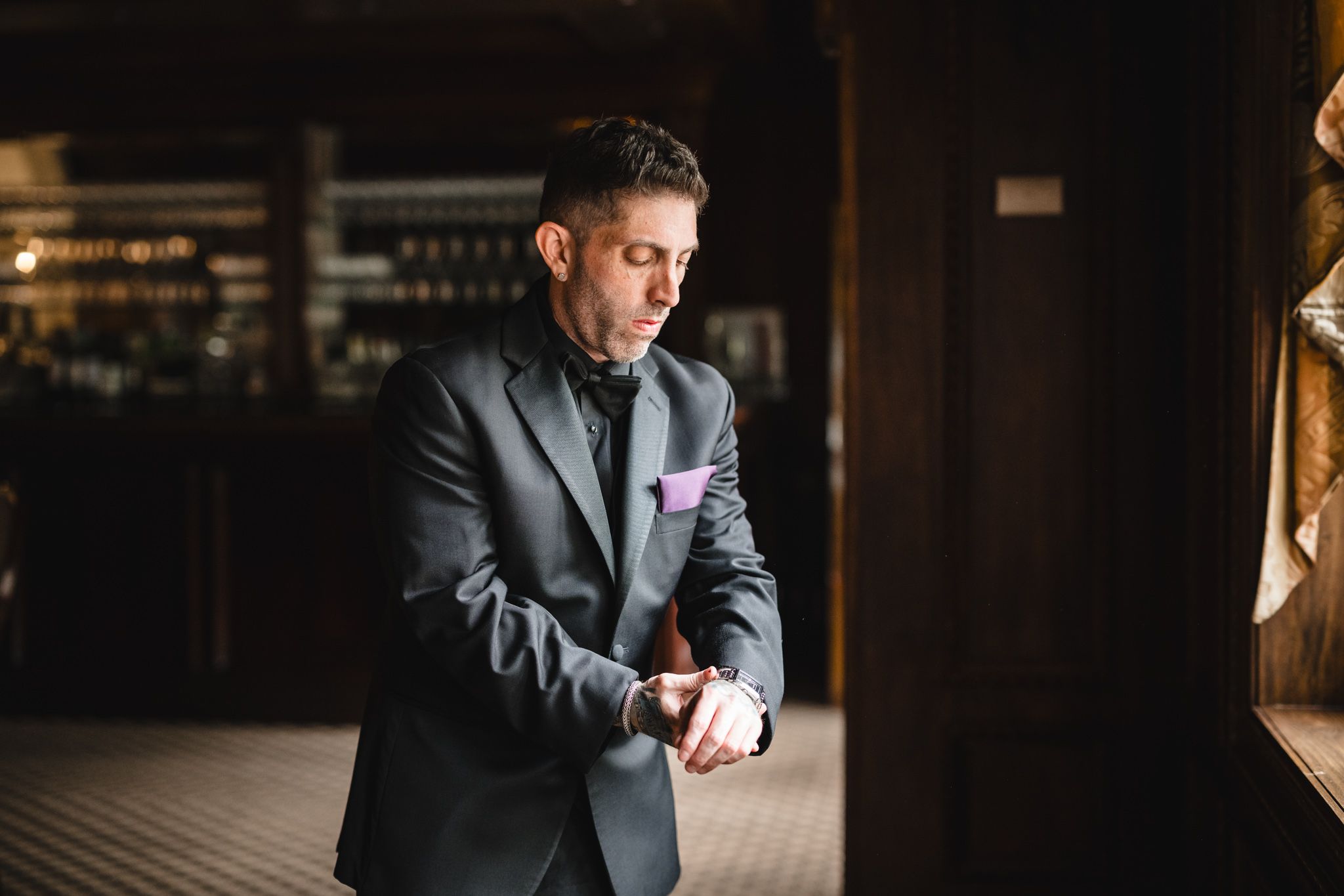 Man in dark suit and purple tie adjusts his cuffs while standing by a window.