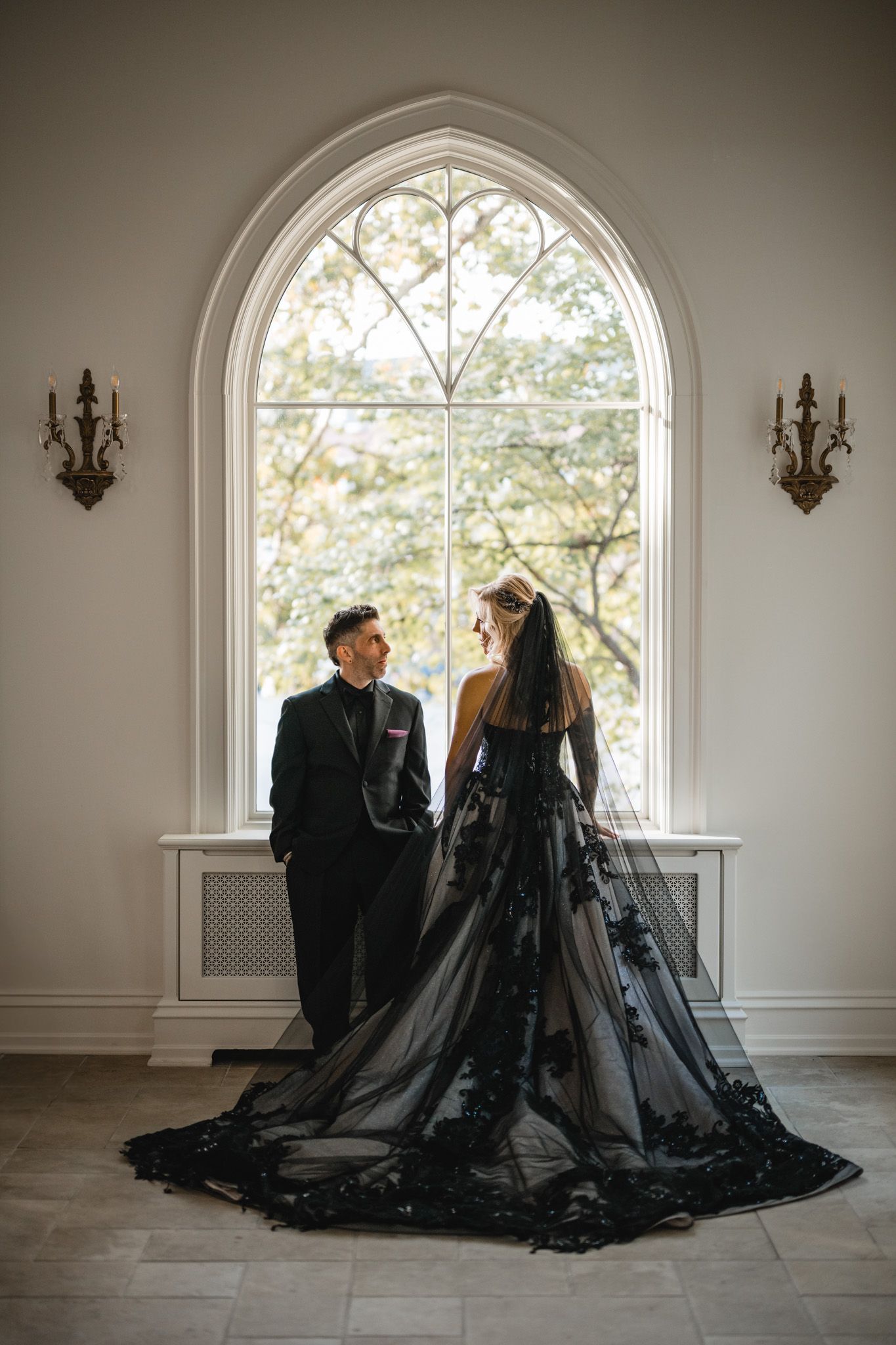 A couple in black formal wear stands beneath a grand arched window with wall sconces.