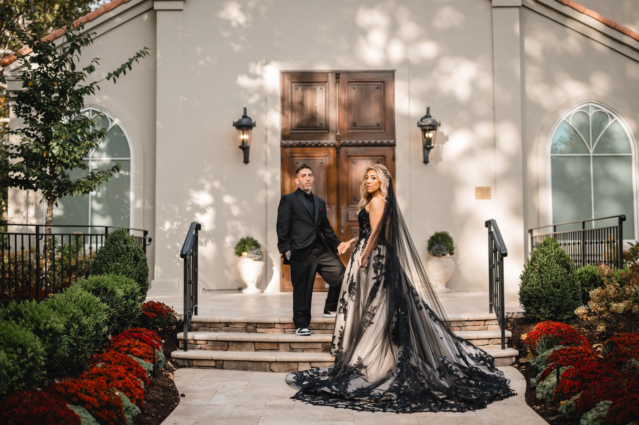 Elegant couple posing outside white church building with red flowers and arched windows.