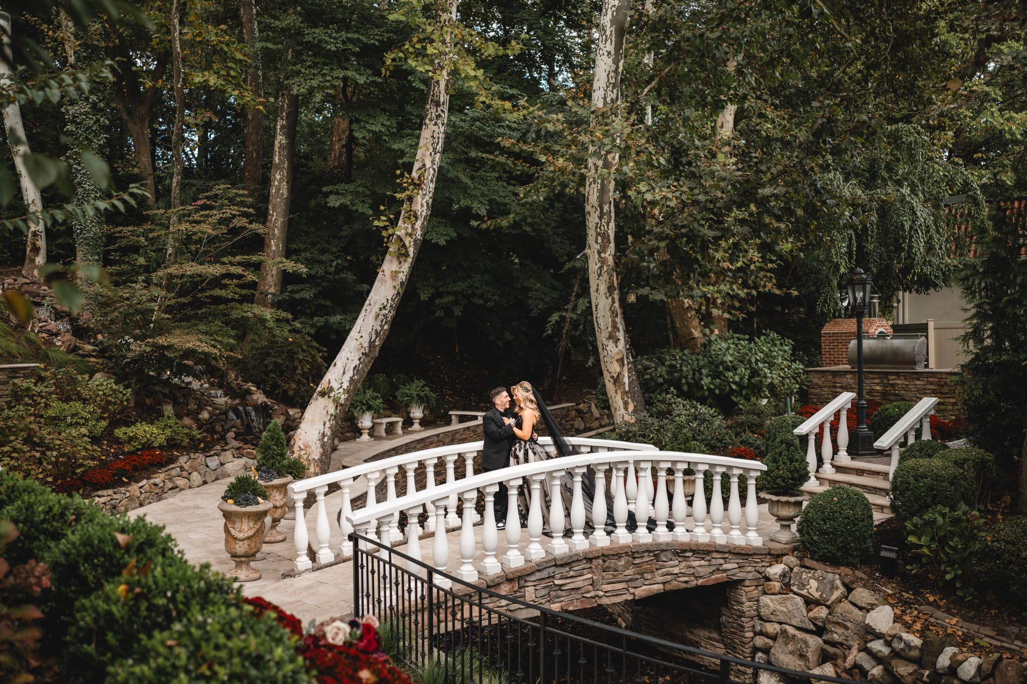 A couple shares a romantic moment on an ornate white garden bridge surrounded by lush trees and greenery.