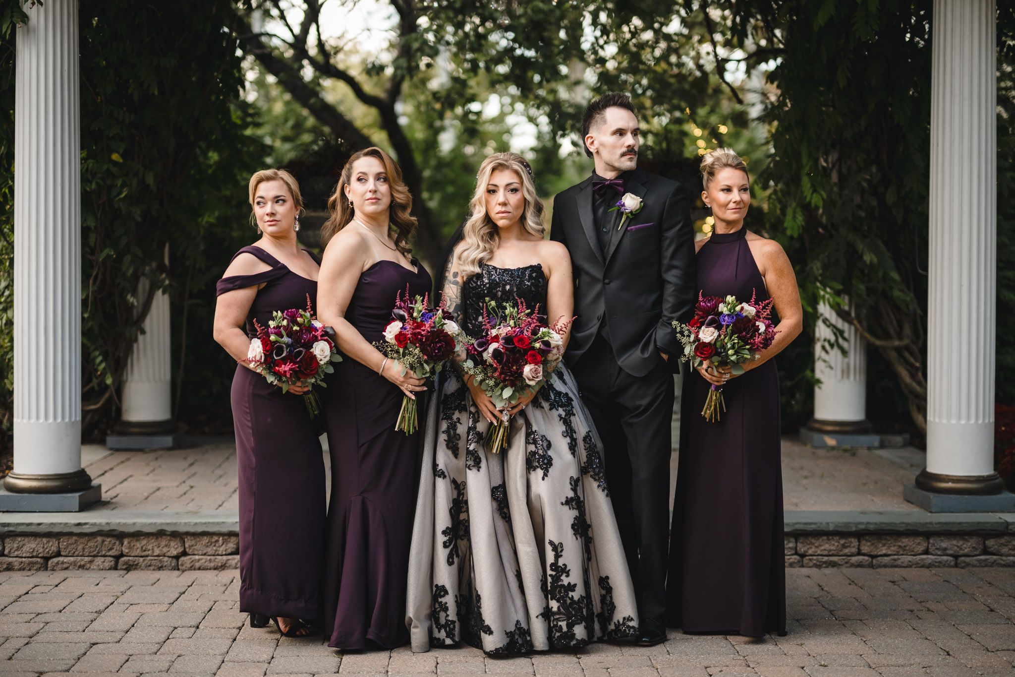 Wedding party in black and purple attire poses between white columns outdoors.
