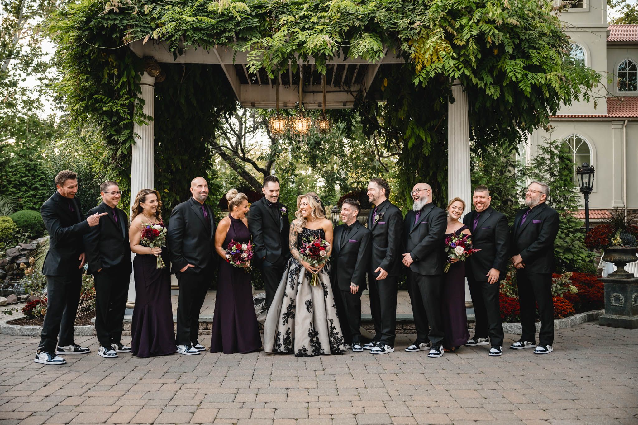 Large wedding party in formal black attire poses under a garden pergola.