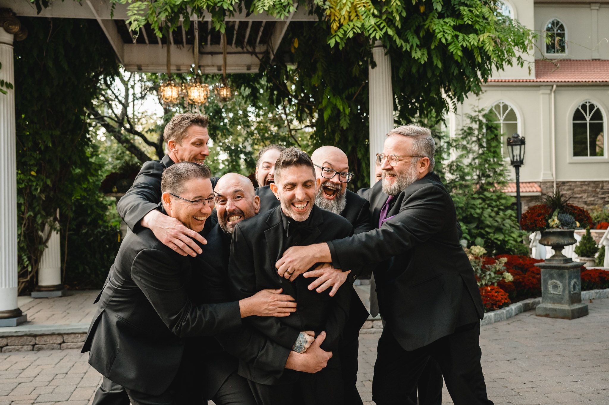 Groomsmen in black suits share a fun group hug moment outdoors.