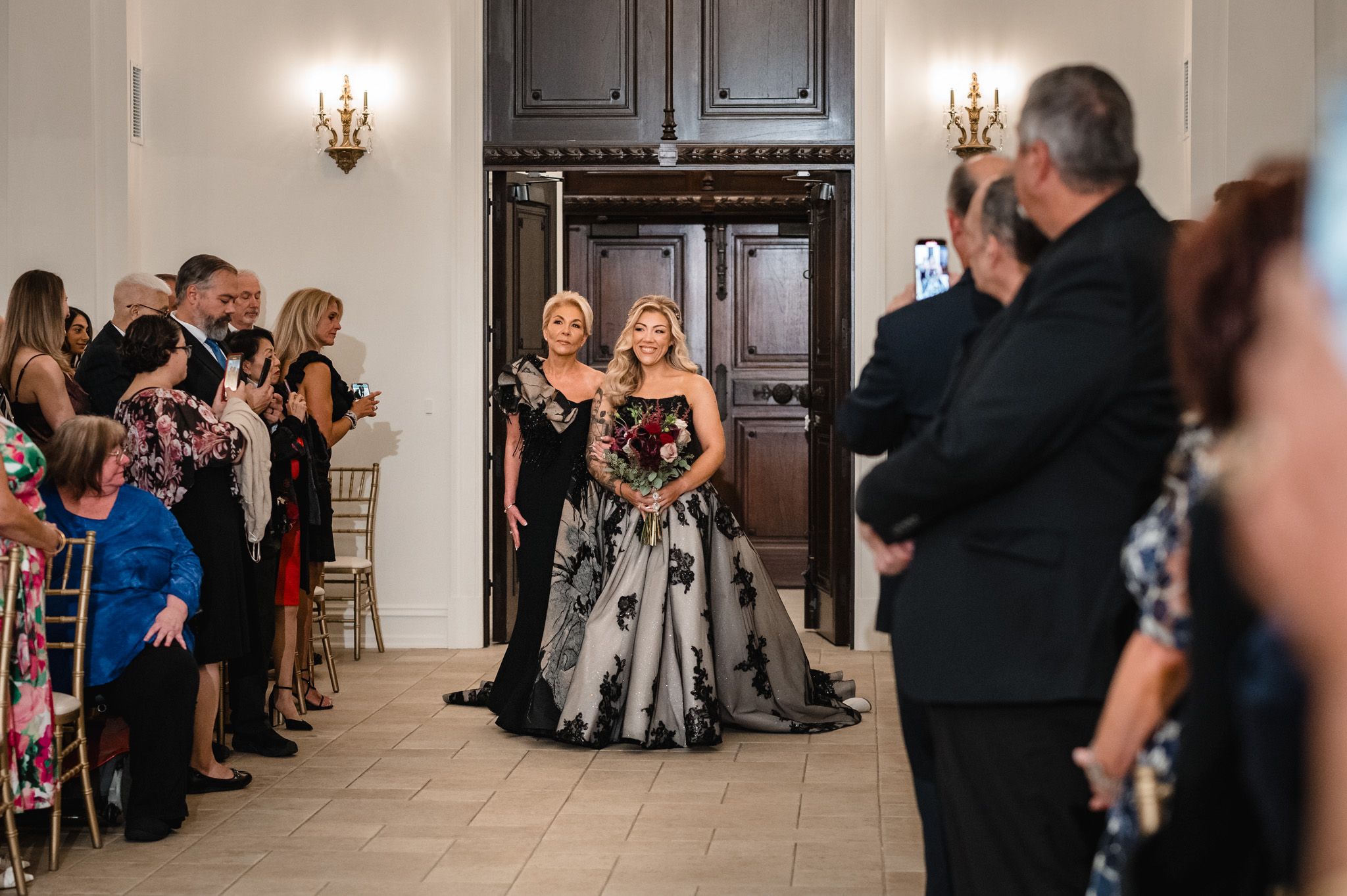 Wedding guests gather in an elegant venue hall with chandeliers and arched windows during a reception celebration.