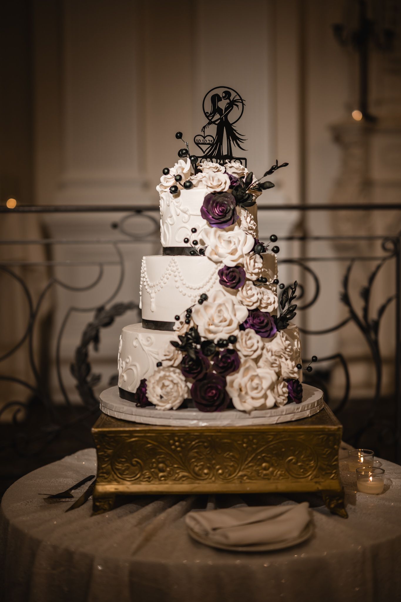 White wedding cake decorated with purple and white roses on a gold stand.