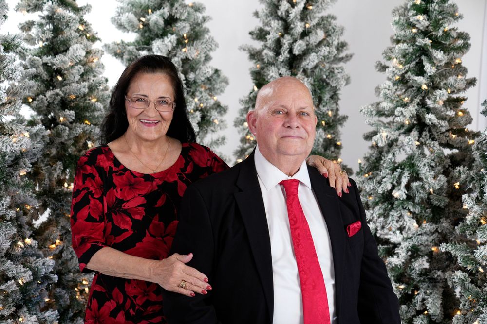 A couple in formal attire poses in front of snow-covered Christmas trees for a holiday photo shoot.