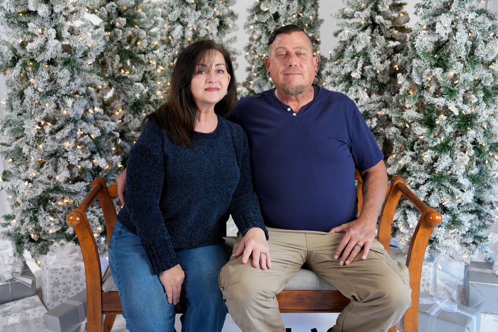 A couple sits together on a wooden bench against a backdrop of snow-covered evergreen trees for a winter portrait.