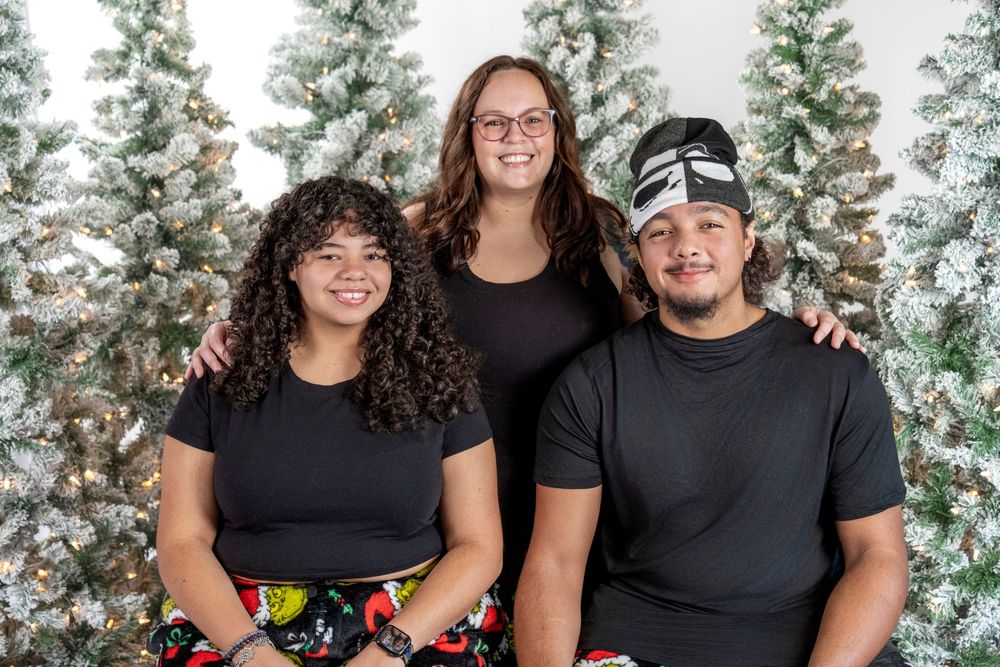 Three people in black t-shirts pose together in front of snowy evergreen trees for a holiday photo.