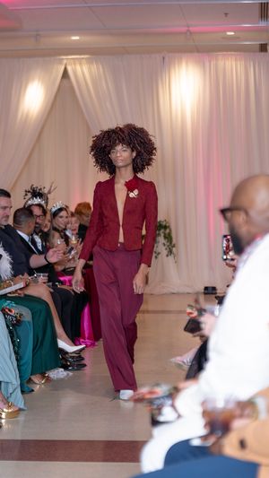 A deep burgundy power suit ensemble being presented on a fashion runway with white backdrop and audience.