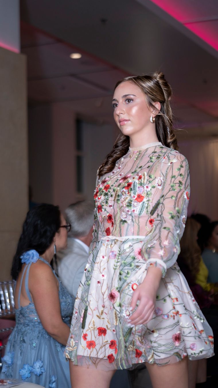 Model walks runway in floral sheer overlay romper with red and pink embellishments during fashion show.