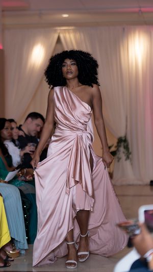 Model walks runway in flowing pink satin gown with dramatic draping at fashion show.