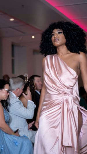 Model wearing a shimmering pink satin wrap dress walks the runway under pink lighting at a fashion show.
