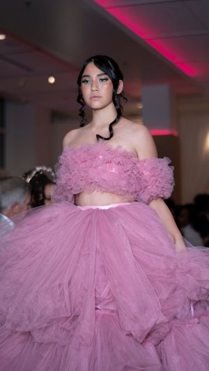 Model walks runway in voluminous pink tulle off-shoulder gown with dramatic ruffled details at fashion show.