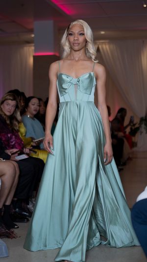 Fashion models present flowing mint green gowns on a runway with dramatic lighting and audience members visible.