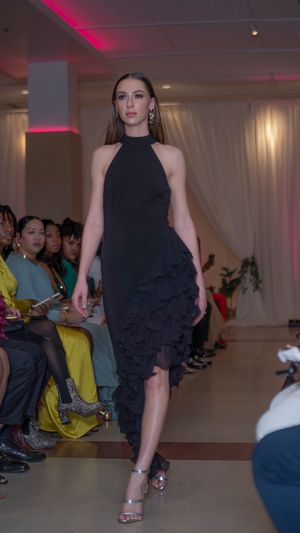 Model walks runway in black ruffled cocktail dress with pink accent lighting.