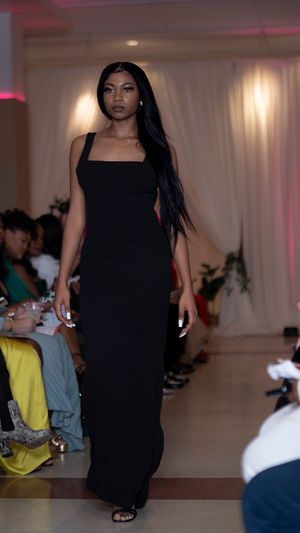 Model showcases flowing black evening gown on illuminated runway with pink accent lighting.