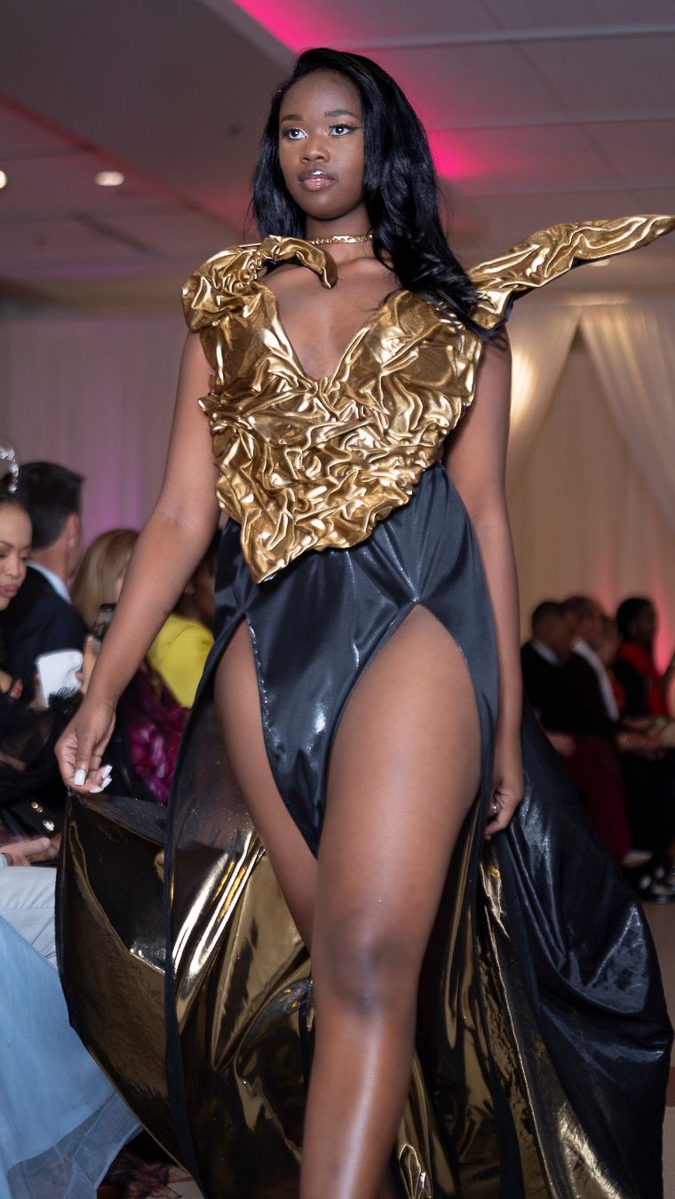 Model struts down catwalk wearing dramatic gold and black bodysuit with metallic details.