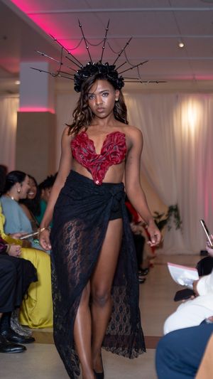 Fashion model walks runway in black lace dress with dramatic red floral shoulder detail.